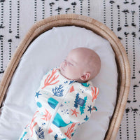 Newborn baby swaddled in a dreamy days dream swaddle ocean themed, lying in a Moses basket with a patterned blanket carpet..