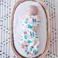 Newborn baby swaddled in a colorful dreamy days dream swaddle with ocean-themed design, lying on a white surface with black text pattern.
