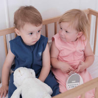 Two toddlers wearing the Dreamy days navy dream swaddle and the pink dream swaddle holding a dream pod white noise machine in cot