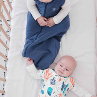 Baby and toddler swaddled in a colourful dreamy days dream swaddle with ocean-themed design, and navy dream sack lying on a white surface.