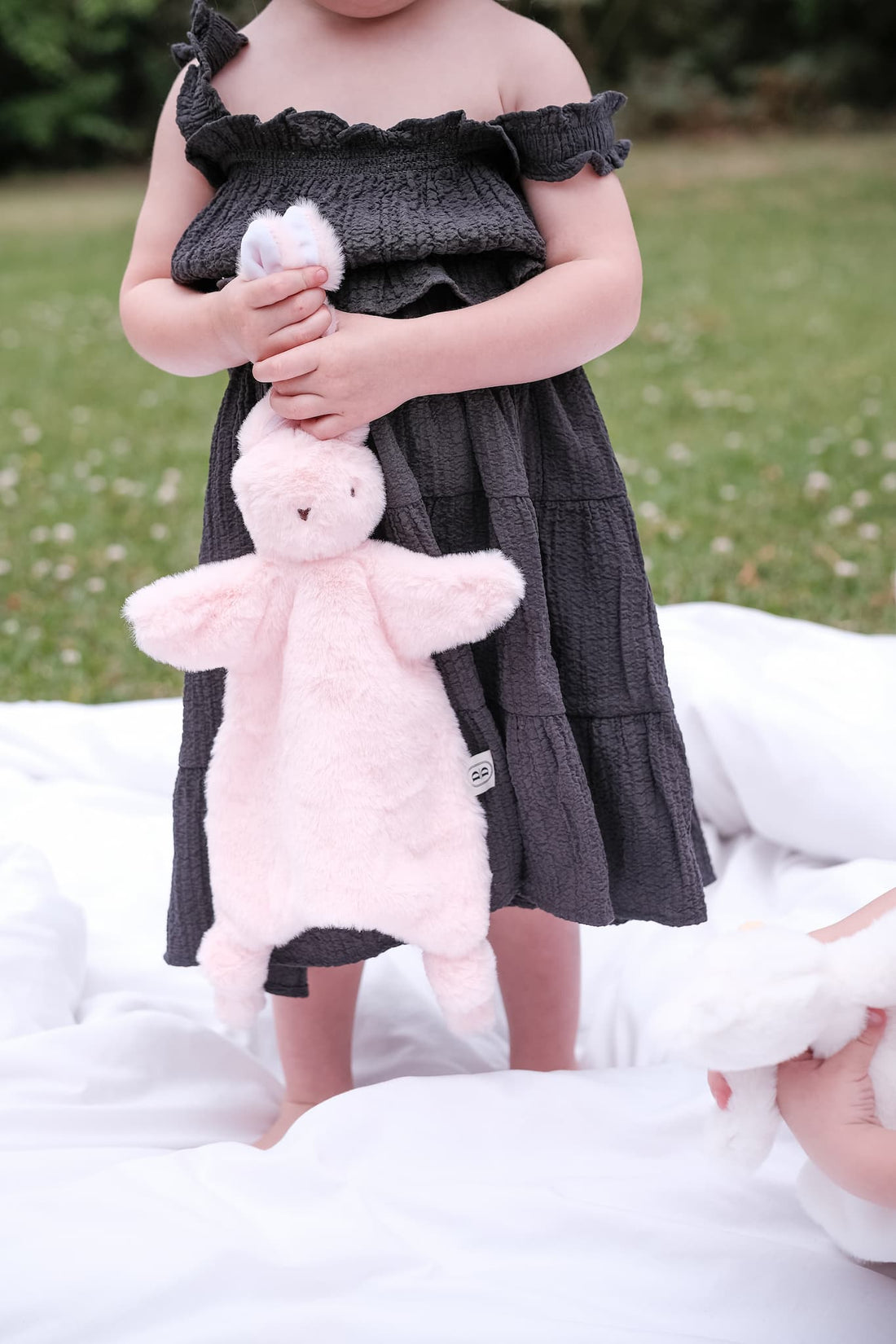 Child in a dark dress holding a pink bunny comforter on a grassy area.