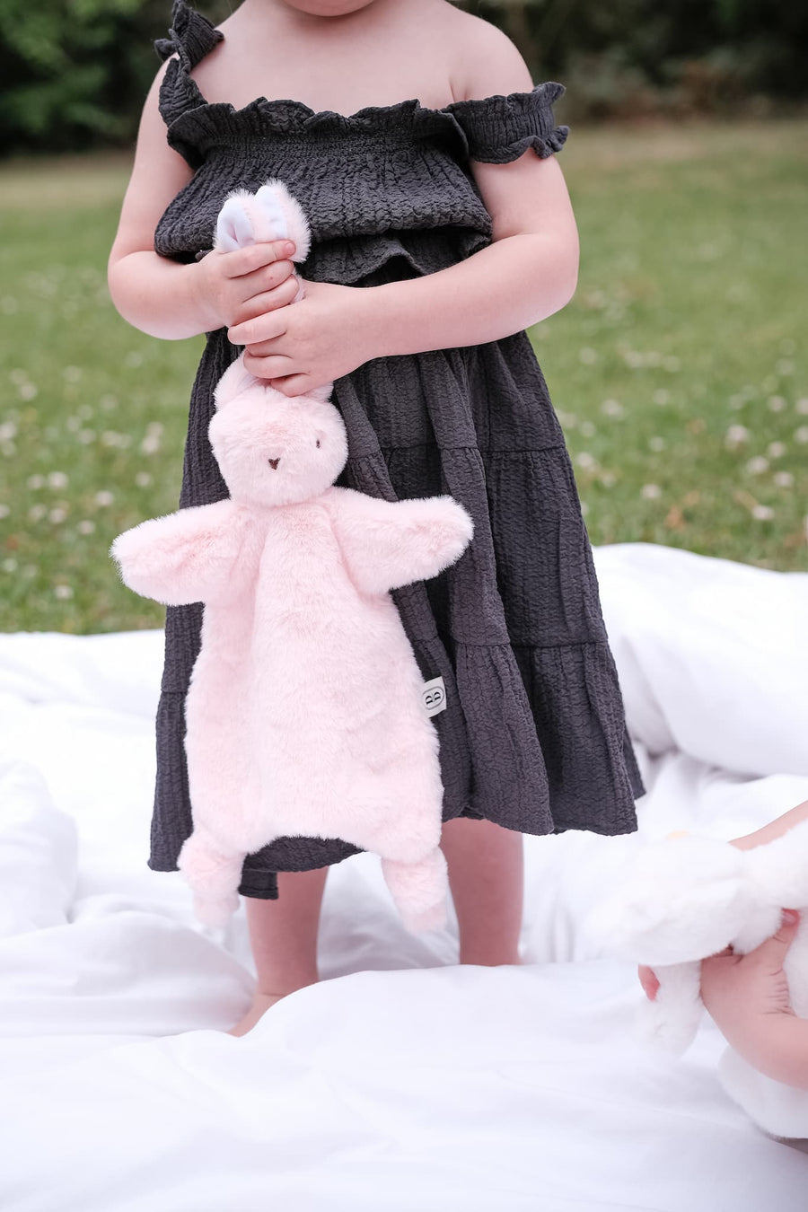 Child in a dark dress holding a pink bunny comforter on a grassy area.