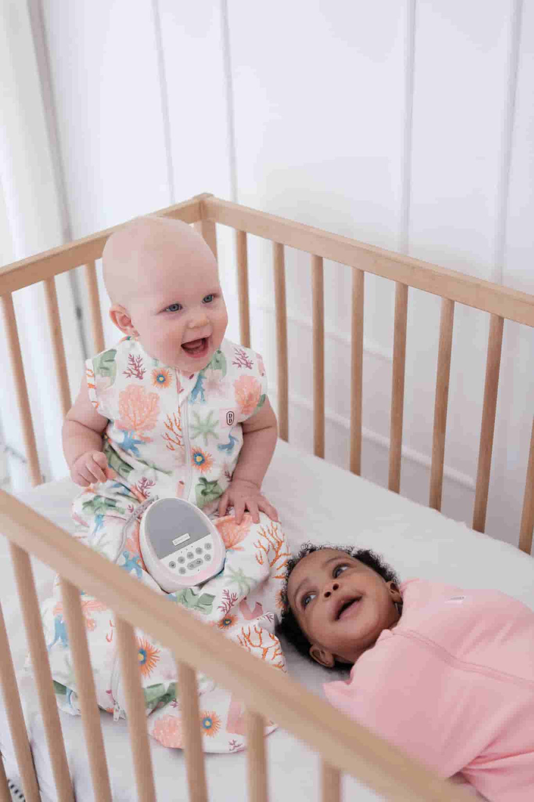 Two babies in a crib wearing the Dreamy Days dream swaddle with one wearing a  pink coral and the other in pink together with the Dream pod white noise machine.