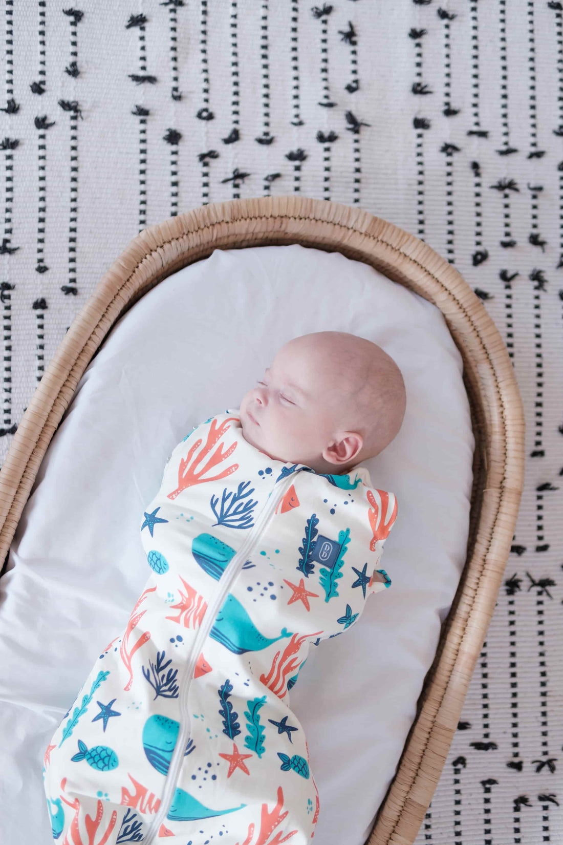 Newborn baby swaddled in a dreamy days dream swaddle ocean themed, lying in a Moses basket with a patterned blanket carpet..