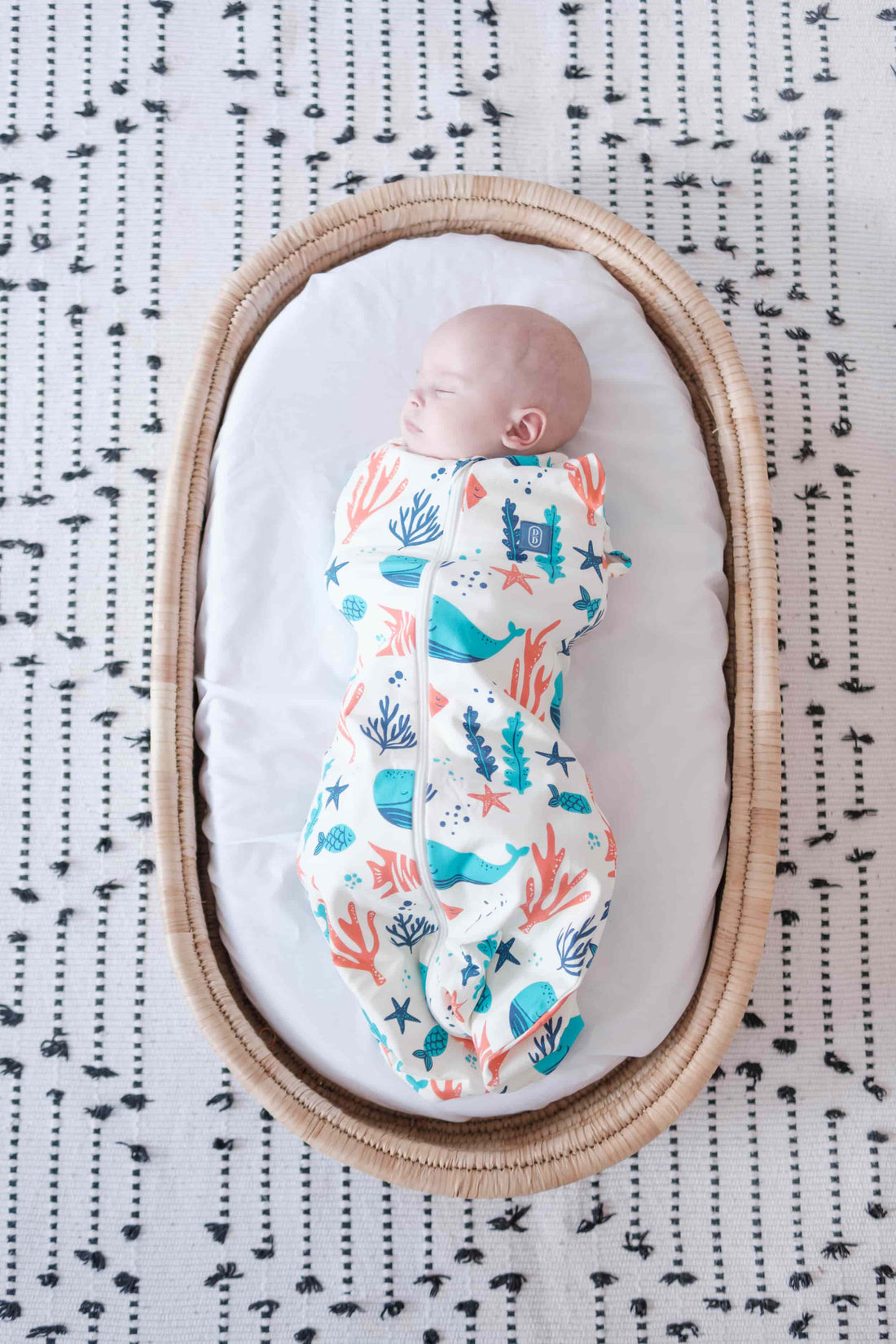 Newborn baby swaddled in a colorful dreamy days dream swaddle with ocean-themed design, lying on a white surface with black text pattern.