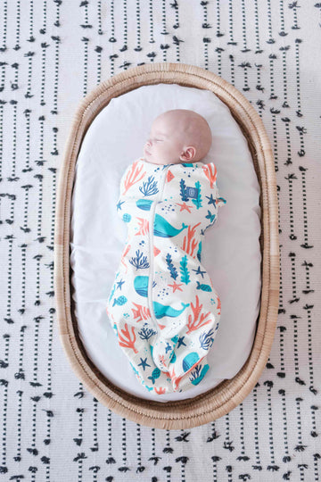 Newborn baby swaddled in a colorful dreamy days dream swaddle with ocean-themed design, lying on a white surface with black text pattern.