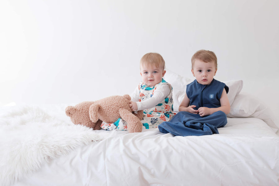 Two toddlers wearing the dreamy days navy and ocean themed dream sacks on white bed with a teddy bear