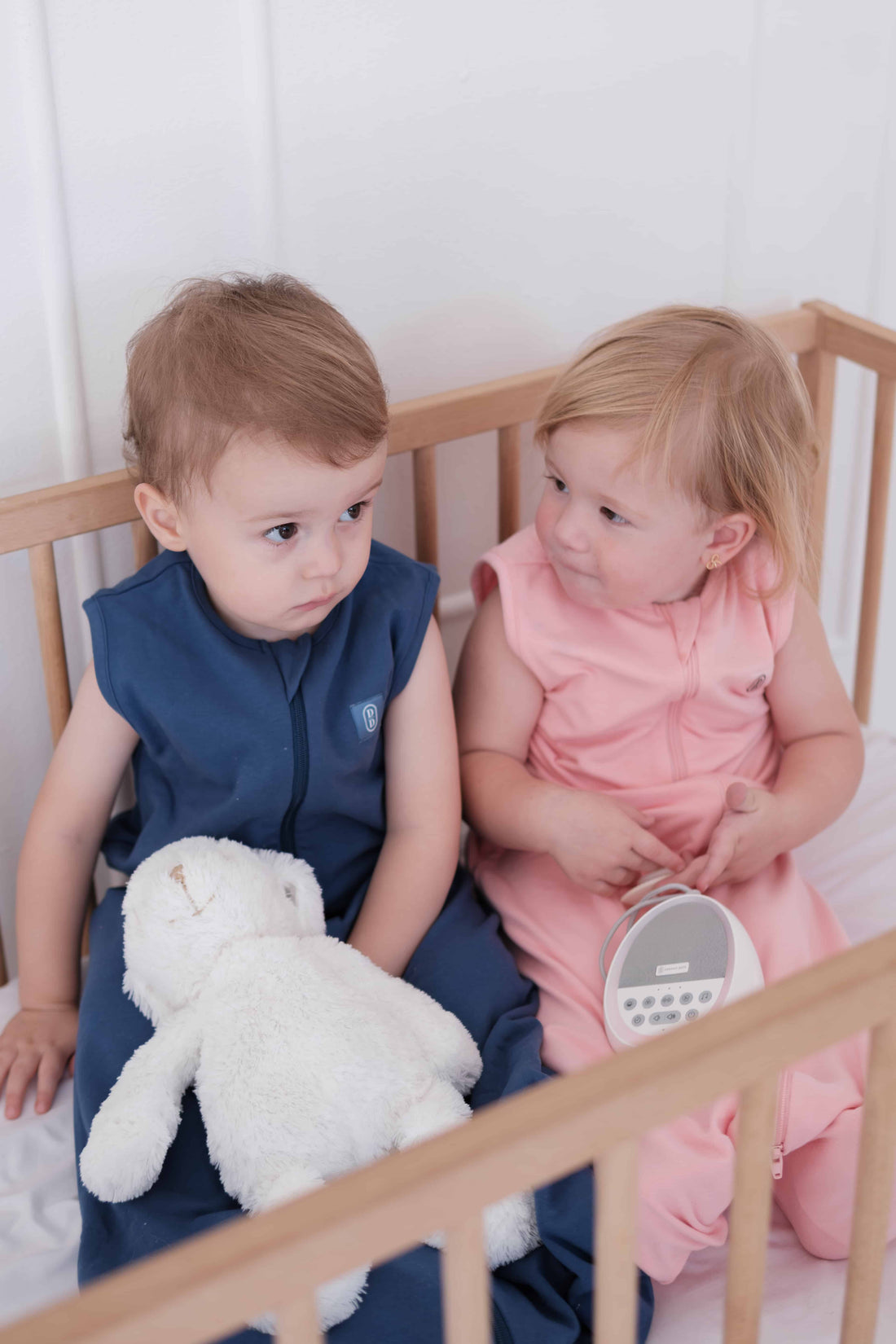 Two toddlers wearing the Dreamy days navy dream swaddle and the pink dream swaddle holding a dream pod white noise machine in cot