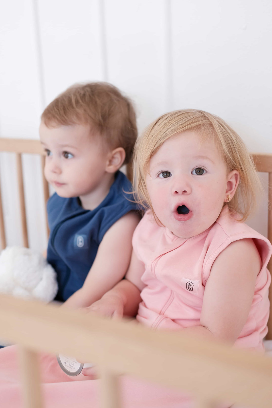 Two toddlers wearing the Dreamy days navy dream swaddle and the pink dream swaddle holding a dream pod white noise machine