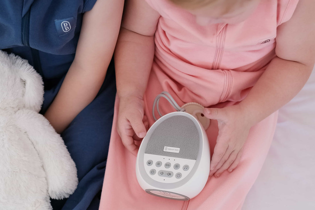 Two toddlers wearing the Dreamy days navy dream swaddle and the pink dream swaddle holding a dream pod white noise machine