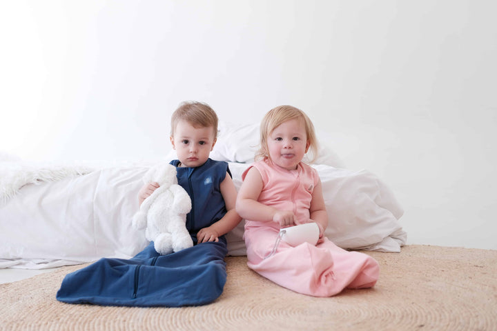 Two toddlers sitting by bed wearing the Dreamy days navy dream swaddle and the pin dream swaddle