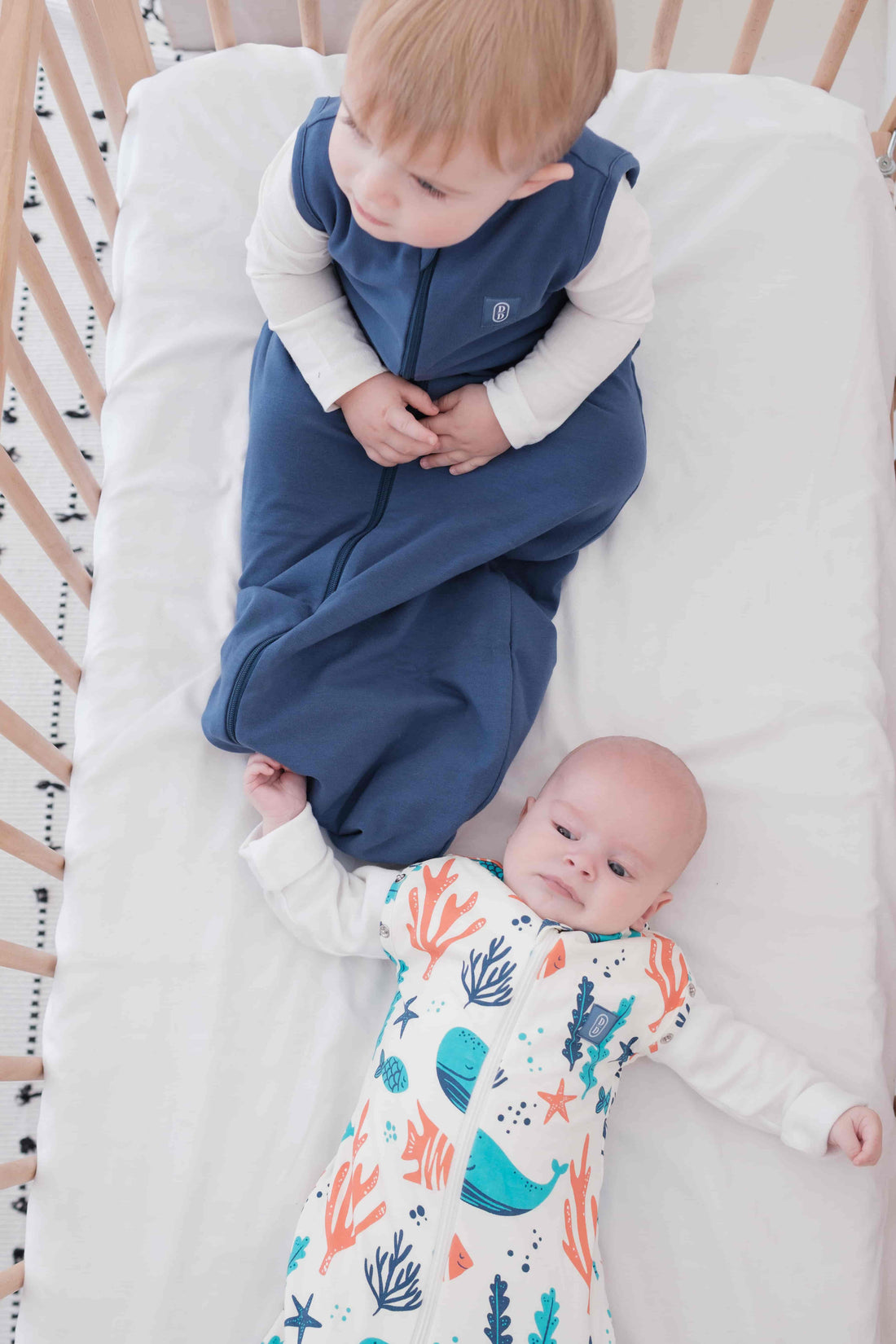 Baby and toddler swaddled in a colourful dreamy days dream swaddle with ocean-themed design, and navy dream sack lying on a white surface.