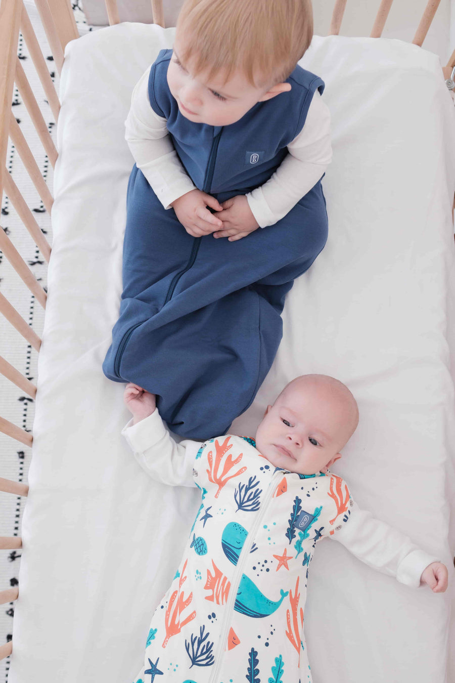 Baby and toddler swaddled in a colourful dreamy days dream swaddle with ocean-themed design, and navy dream sack lying on a white surface.